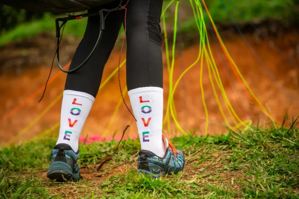 Mädchenbeine auf einer grünen Startwiese, im Hintergrund ein bereitliegender Gleitschirm, weisse Socken mit dem Schriftzug „LOVE“ in bunten Buchstaben.
